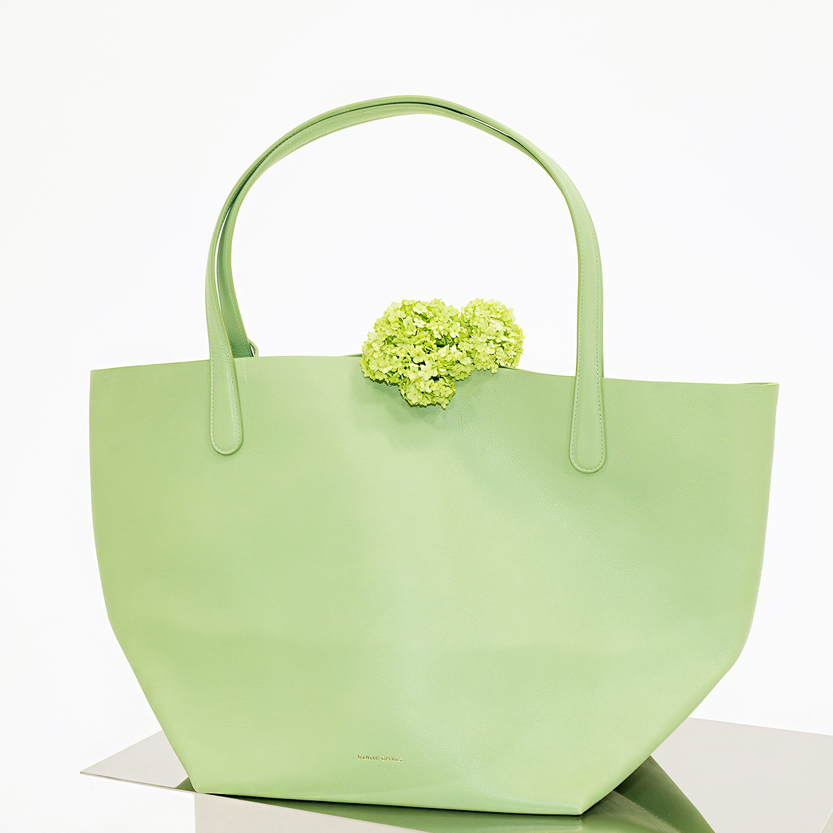 WWD shopping editors test out the Mansur Gavriel Everyday Soft Tote in light green colorway on silver display table with floral accent on bag